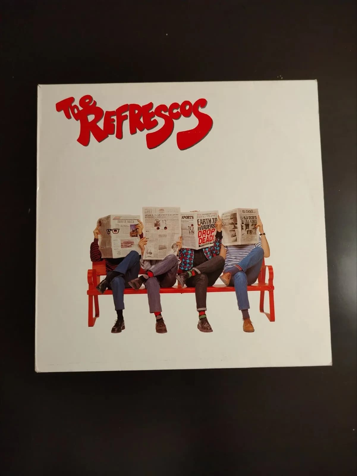 Portada de álbum The Refrescos con cuatro personas en un banco leyendo periódicos.