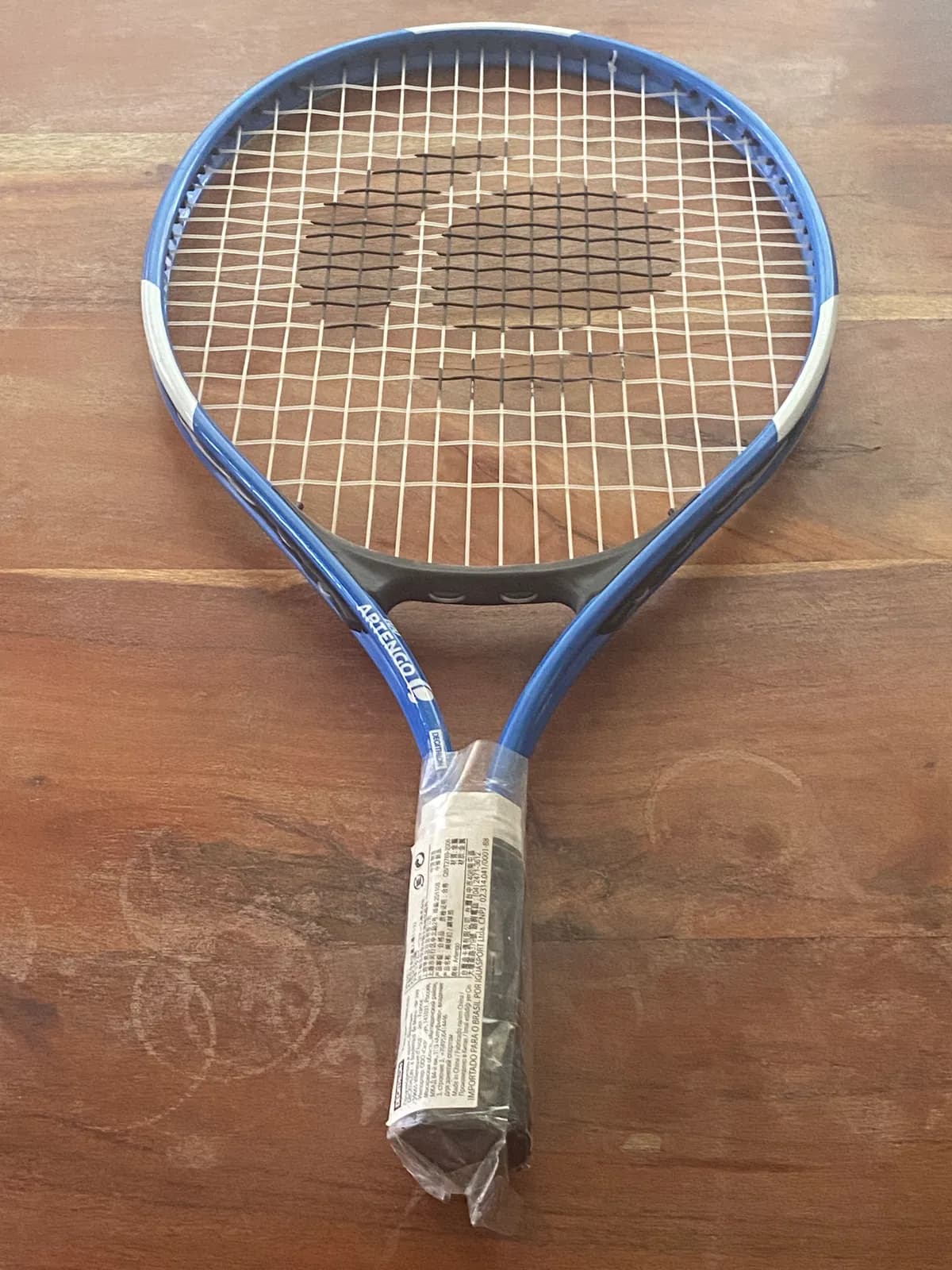 Raqueta de tenis Artengo azul y blanco, con mango envuelto en plástico protector, sobre madera.