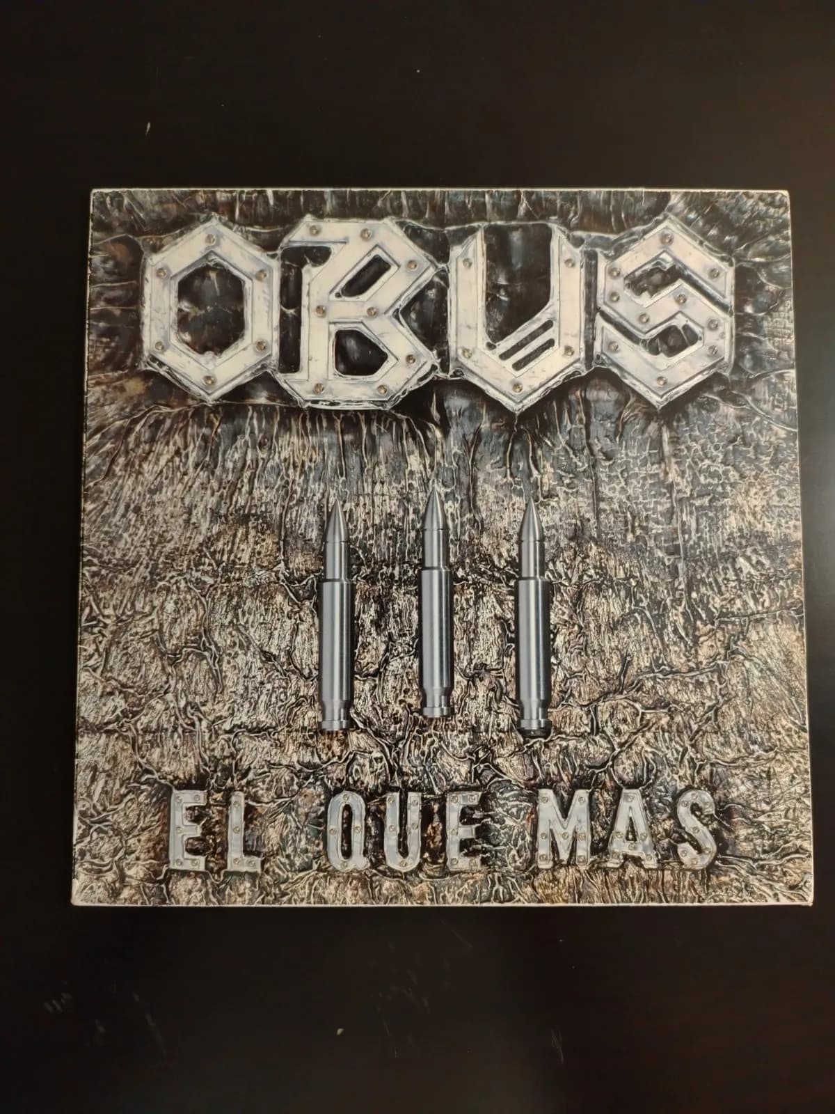 Portada de disco de vinilo Obús El Que Más con letras metálicas, balas y textura rugosa.